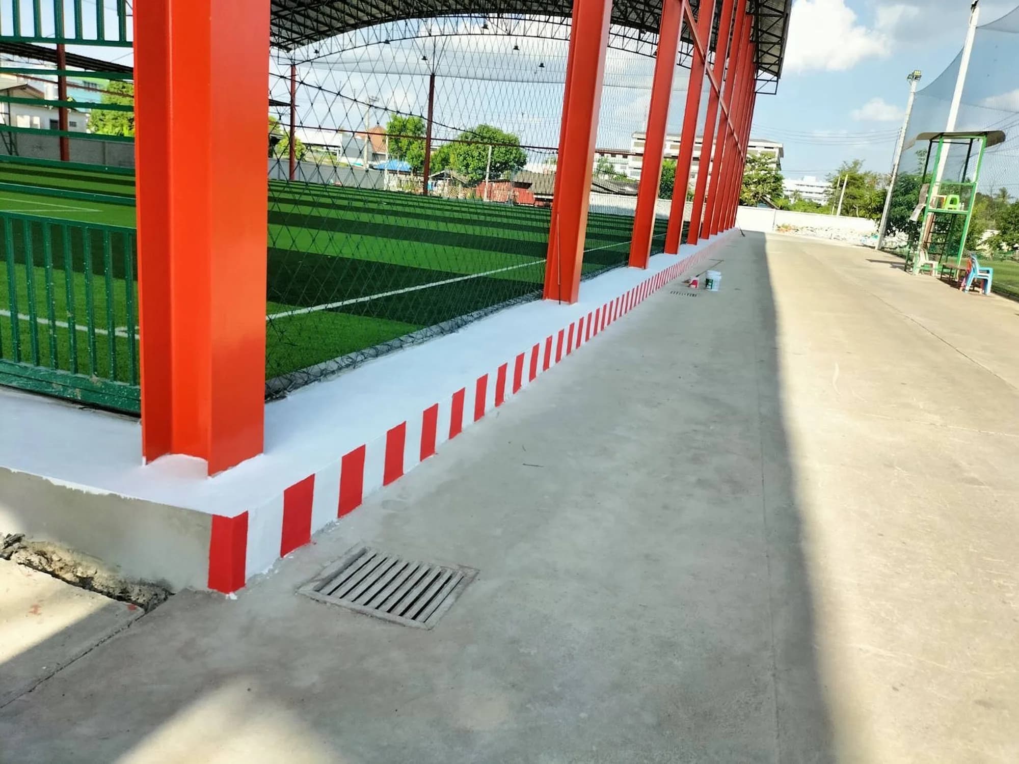 ขอบปูนทาสีขาวแดงตามแนวเสาเหล็กสีส้ม ข้างสนามหญ้าเทียมและทางเดินคอนกรีต
