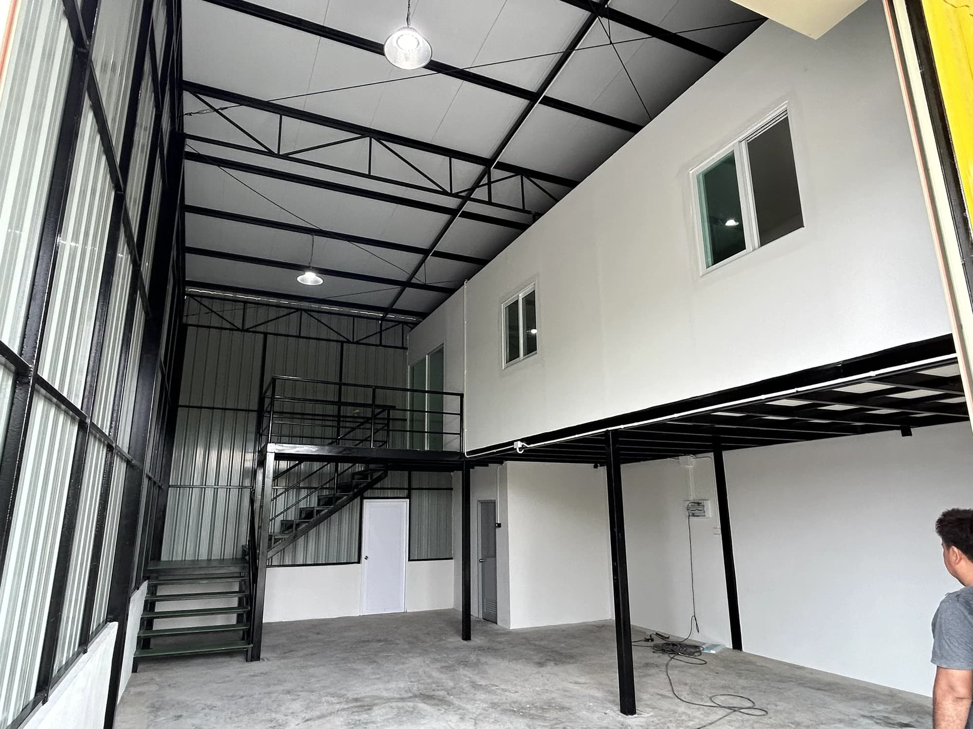 ภายในโรงงานมีชั้นลอยผนังขาวพร้อมหน้าต่างกระจก บันไดเหล็กสีดำ และโครงหลังคาเหล็กเปลือย