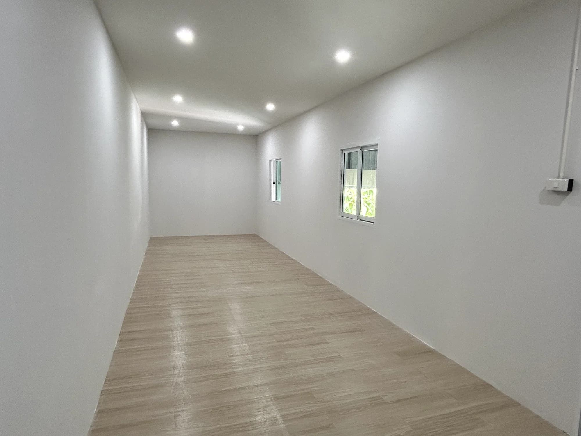 ห้องภายในผนังขาว พื้นลายไม้สีอ่อน ไฟดาวน์ไลท์บนเพดาน และหน้าต่างบานเลื่อนมองออกนอกอาคาร