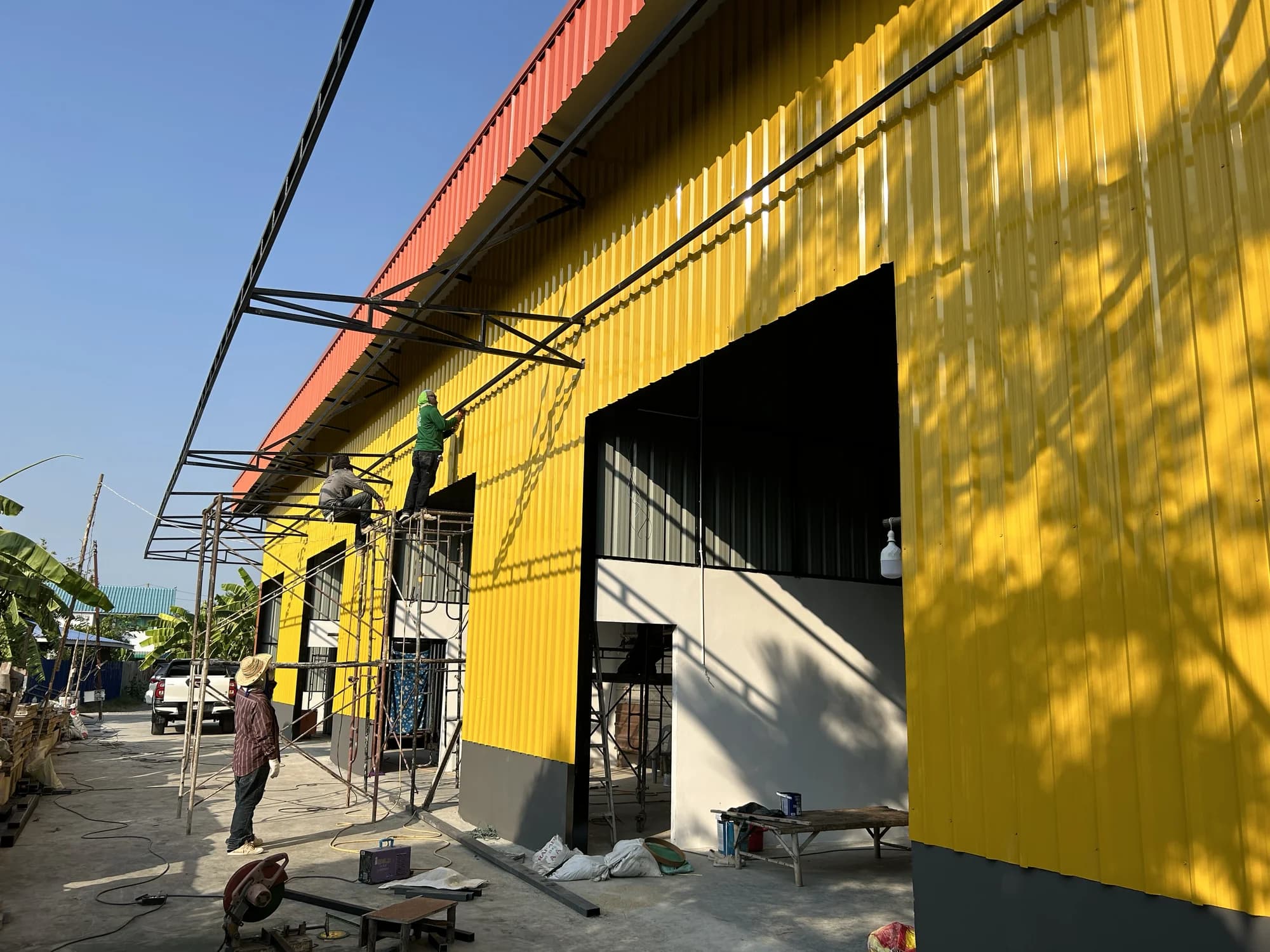 ช่างบนนั่งร้านกำลังติดตั้งโครงเหล็กกันสาดกับผนังเมทัลชีทสีเหลือง มีเครื่องมือและวัสดุกระจายบนพื้นคอนกรีต