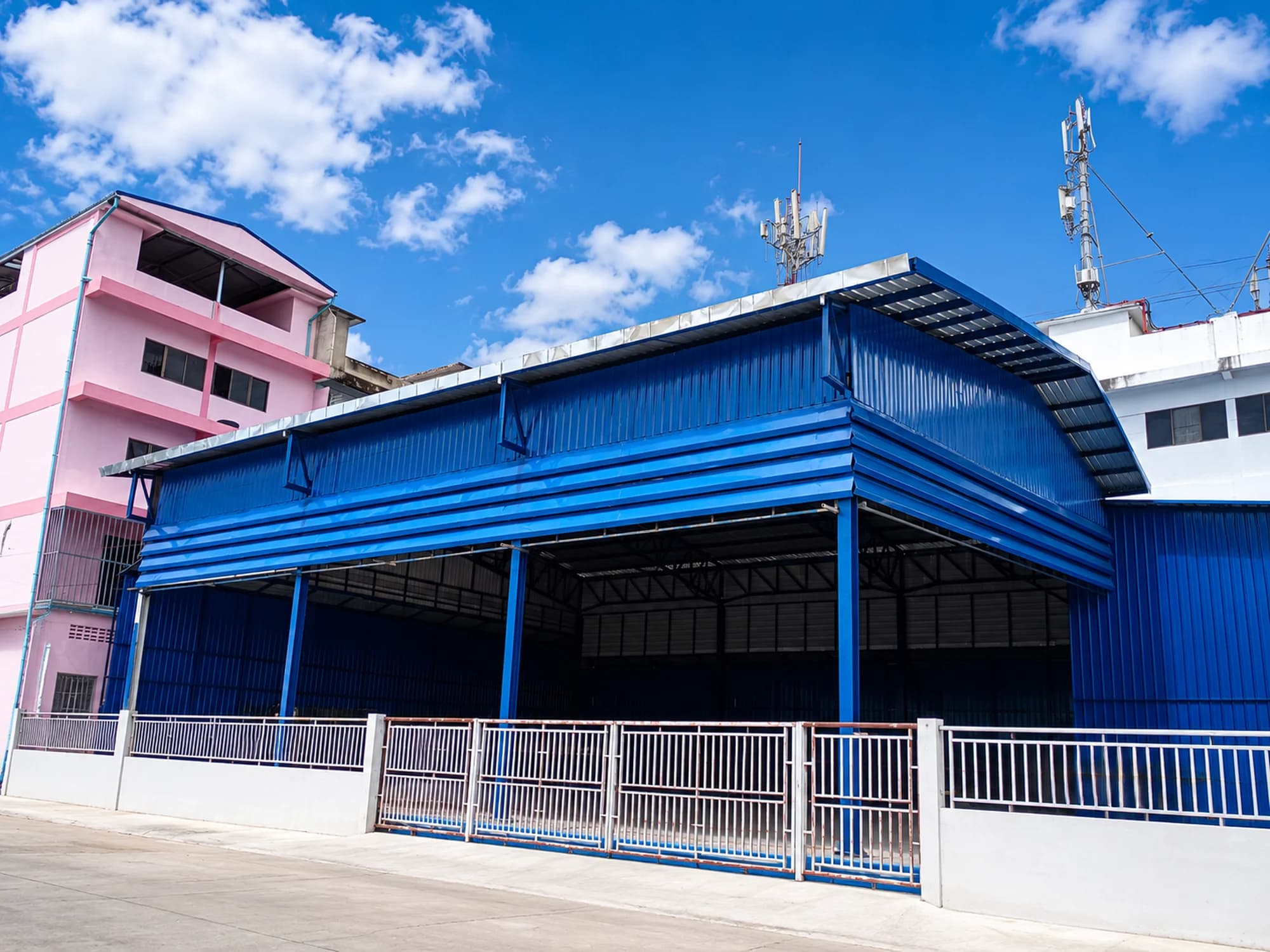 อาคารโรงงานผนังและหลังคาเมทัลชีทสีน้ำเงิน ช่องเปิดด้านหน้าโล่ง มีรั้วเหล็กและประตูเลื่อนหน้าลาน
