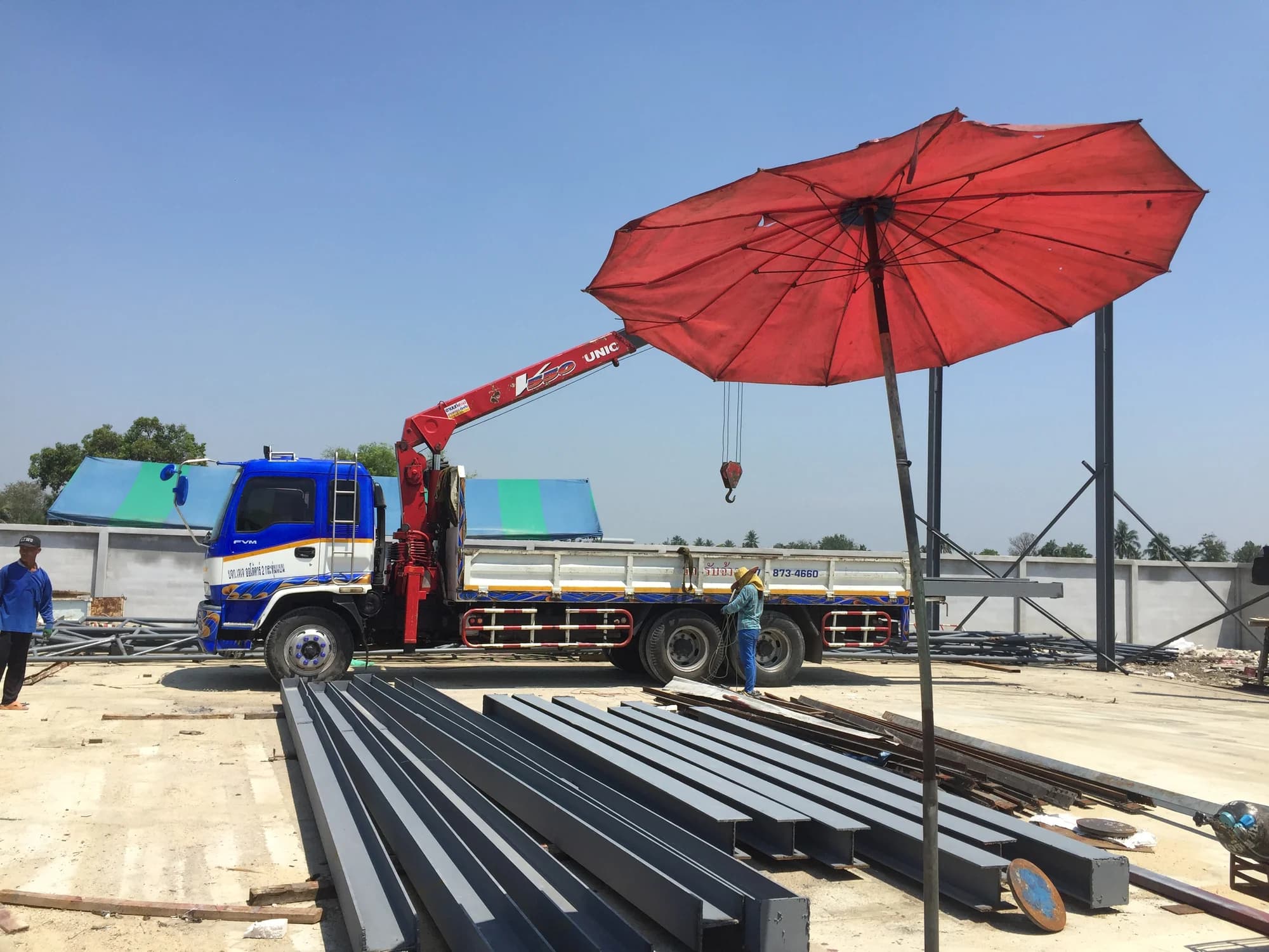 รถบรรทุกติดเครนสีฟ้าแดงจอดบนพื้นคอนกรีตข้างกองคานเหล็กตัวไอสีเทา มีคนงานยืนแถวโครงสร้าง